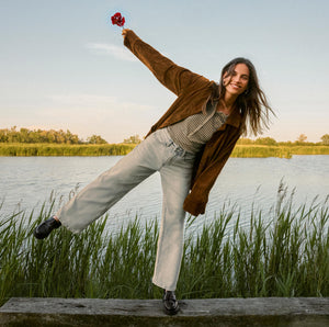  Woman in a brown jacket and light jeans standing on a wooden bench by a lake, holding a red flower.