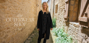  Woman walking down a narrow stone alleyway wearing a black coat with 'the Outerwear Shop' branding.