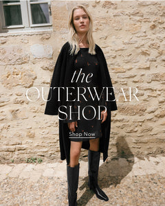  Woman wearing a black coat and dress with text overlay 'the OUTERWEAR SHOP Shop Now' against a stone wall.