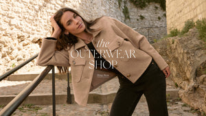  Woman wearing a beige coat with text overlay 'the OUTERWEAR SHOP' against a stone wall background.