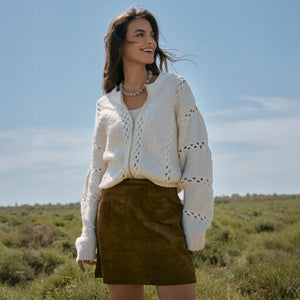 DressesMelbourne Sandy Bay Palm Midi Dress Woman wearing a white knitted cardigan and brown skirt standing in a field with a blue sky.