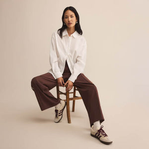 Person sitting on a stool wearing a white shirt and brown pants against a beige background