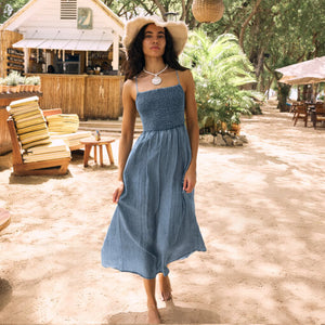 SweatersLooking Glass Crochet Polo Sweater Woman in a blue dress and hat standing in a tropical setting with wooden furniture and plants.