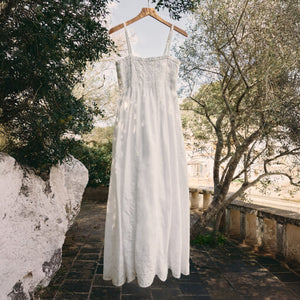  White dress hanging on a wooden hanger against a natural outdoor background with trees and stone wall.