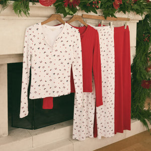  White long-sleeve shirt with red and black pattern, red pants, and white skirt with red and black pattern hanging on a wooden hanger against a decorated fireplace.