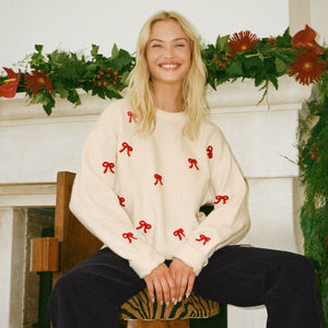  Woman wearing a cream sweater with red bows sitting in front of a decorated fireplace.