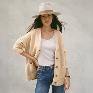 Woman wearing a beige cardigan, white top, blue jeans, and a beige hat against a plain background