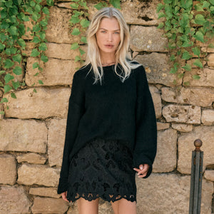  Woman wearing a black sweater and black lace skirt against a stone wall with greenery.