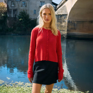  Woman in a red sweater and black skirt standing by a body of water with a stone bridge in the background.
