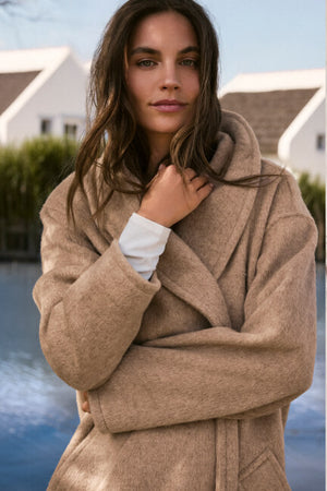 Woman wearing a beige coat standing by a body of water with houses in the background