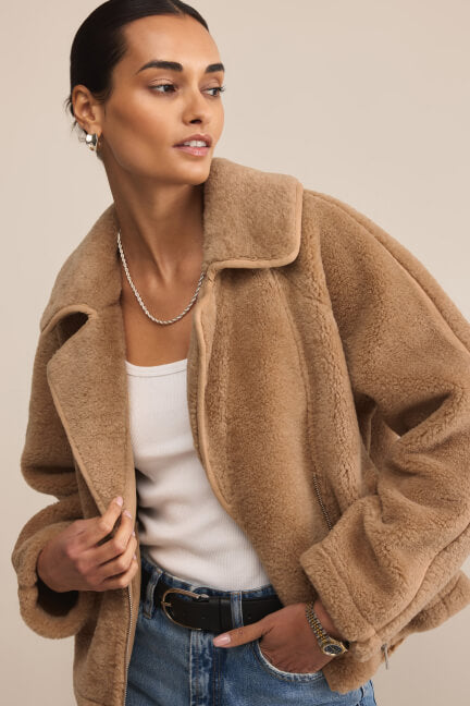Woman wearing a beige teddy coat over a white top and blue jeans on a plain background