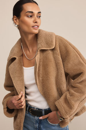 Woman wearing a beige teddy coat over a white top and blue jeans on a plain background