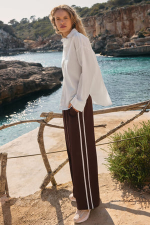 Woman in a white blouse and brown pants with white stripes standing by a scenic coastal area.