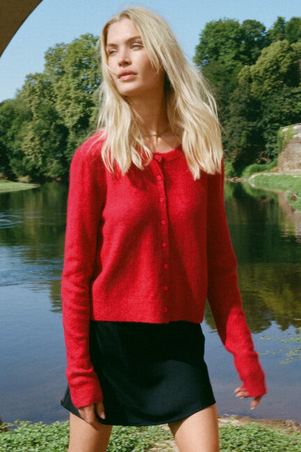 Woman wearing a red sweater and black skirt standing by a body of water with trees in the background