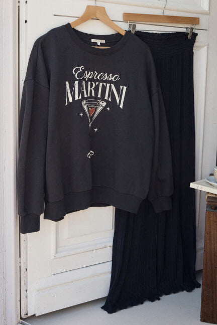 Black sweatshirt with 'Espresso Martini' design and black skirt hanging on a wooden hanger against a white door.
