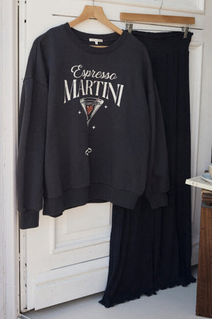Black sweatshirt with 'Espresso Martini' design and black skirt hanging on a wooden hanger against a white door.